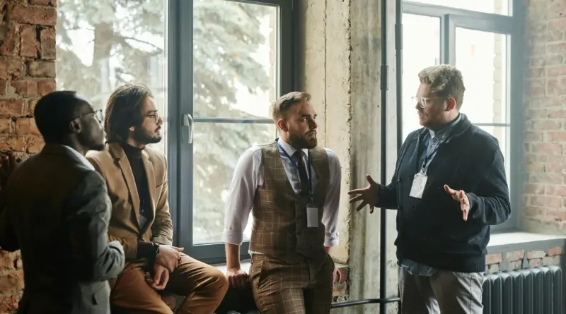 Cuatro profesionales de negocios manteniendo una conversación en un pasillo industrial junto a un ventanal, donde tres de ellos adoptan posturas de evaluación analítica frente a un intermediario que gesticula activamente, representando el filtro de los family offices ante el brokerage oportunista.