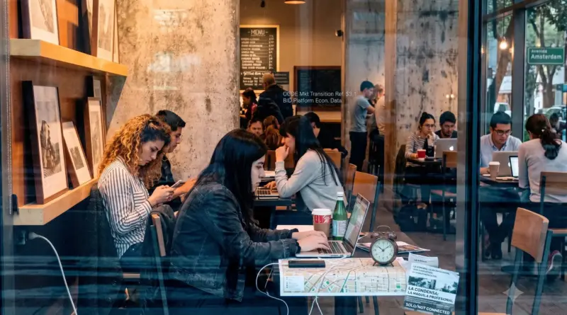 Profesionales independientes trabajando con laptops dentro de una cafetería de retail, visto a través del reflejo del cristal exterior en una zona corporativa urbana.